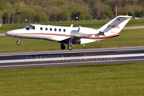 Side view of a white, twin engined biz-jet with the engines mounted on the rear fuselage, flying from right to left at a very low altitude, it's main wheels just a couple of metres above the black runway.
The plane has flaps slightly extended behind the wings, and the nose raised significantly, suggesting it has just taken off.
There is a thin red and black stripe running along the body, and the registration "D-IOHL" on the tail in red with a black drop shadow.
Green grass fills the foreground, with more grass in the background leading up to trees stretching across the frame in the distance.
