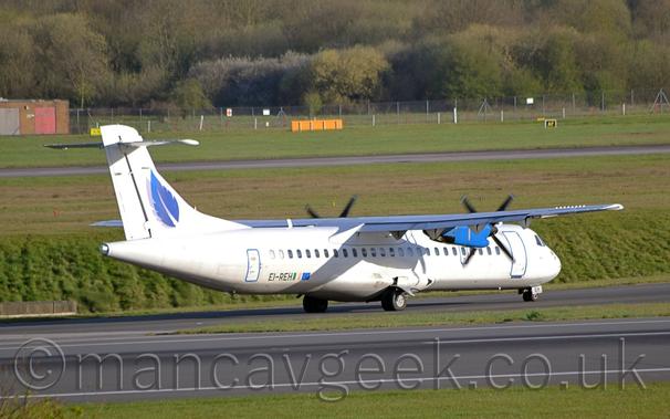 Side view of a white, high-winged, twin propellor-engined airliner taxiing from left to right on a black taxiway.
The plane has a blue leaf with a grey drop-shadow on the tail, the registration "EI-DEH" on the lower rear fuselage next to Irish and EU flags, and blue engine pods under the wings.
A strip of green grass stretches across the bottom of the frame, with another strip of grass separating the runway and taxiway.
More green grass in the background lead up to trees stretching across the frame in the background.