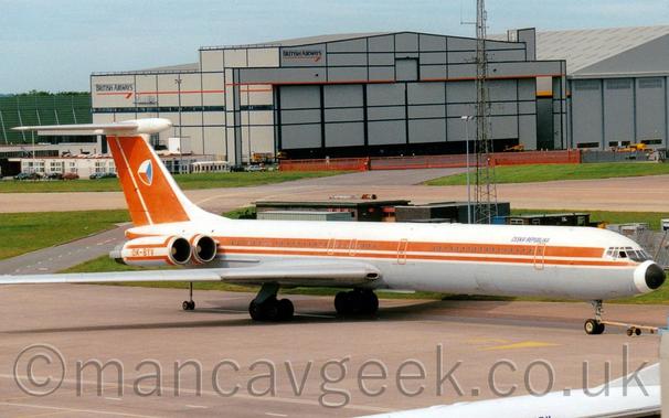 Side view of a white, 4 engined jet airliner with the engines mounted on the sides of the rear fuselage, facing to the right but being pushed back to the left by a low-slung blue truck almost hidden behind a grey building on the left of the frame.
There are faded red stripes running along the body, with red stripes on the white engine pods above and below the red registration "OK-BYV", and a red tail with rounded off cube with white, red, and blue faces.
There are small, black "Ceska Republica" on the upper forward fuselage.
A black pole with wheels at the bottom supports the rear fuselage.
Grey apron fills the bottom of the screen, with the top of a white jet airliner and part of a building in the bottom right corner.
Strips of green grass mark the edges of a taxiway in the background, with a couple of large grey hangars in the distance, under cloudy grey skies.
