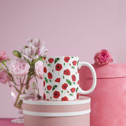 Taza blanca con un diseño de amapolas que he dibujado en acuarela. La taza está encima de una caja redonda y detrás hay un ramo de flores que acompaña al conjunto, todo en tonos claros y pastel rojos y rosas.