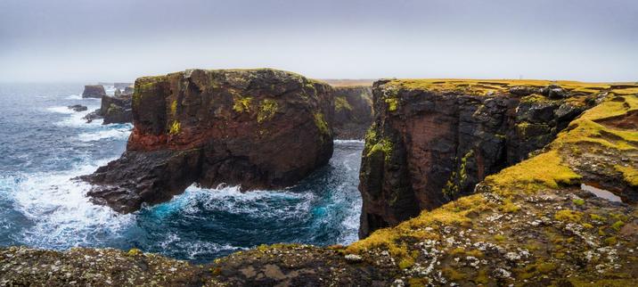 A view of the coast near Eshaness: steep, rocky cliffs plunging into the wavy sea. Yellow-green grasses cover the top of the cliffs as the waves crash onto the rocks below. The sky is dark and gloomy, in contrast with the vibrant blue sea.