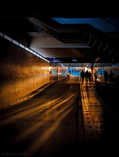 A low sun is shining down into the tunnel for cyclists and pedestrians that runs under Hilversum railway station. The light casts a warm, orange glow on the brick wall and on the ground below. The setting sun also creates a strong, long shadows of the people as they go through the passage.