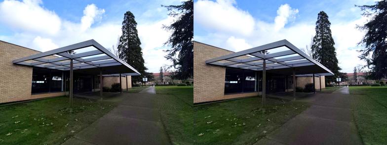 Stereoscopic, side-by-side view. Exterior of the entrance of the Mt. Angel Abbey library. Yellow bricks, with a mid-century modern pergola above the doors. This side of the building is on the top of a hill, so we only see one story.