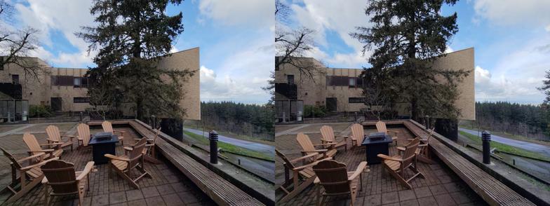Stereoscopic, side-by-side view. Exterior of the Mt. Angel Abbey library. We are looking across a deck full of Adirondack chairs towards a large fir tree, with the side of the building beyond. The building descends the hill from the left side of the picture. We can see two stories across most of the image, with a little bit of the lowest, basement level, on the right. Mostly yellow bricks, with brown, wooden window frames.
