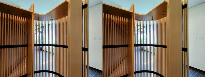 Stereoscopic, side-by-side view. Looking at the inside of a divider in the lobby of the Mt. Angel Library. It is made of thin, vertical blonde wood slats, about one inch thick, separated by similar-sized gaps. It has an undulating shape, curving inward at the ends. (We are looking at the concave side.) The slats are supported by a black steel frame. On the underside of the "lid" of the structure are some coat hooks.