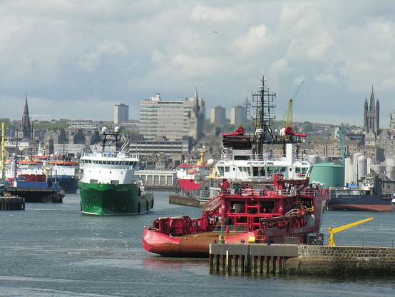 Aberdeen. The image shows a busy scene of ships beyond a breakwater in the bottom right of the frame. A red-hulled one is going away from us and a green-hulled one coming towards us. Others are moored at wharves across the centre of the frame. In the background are buildings, punctuated by high rise blocks and old church and other spires and towers.