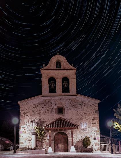 The image captures a traditional stone church at night with a striking star trail effect in the dark sky above. The church features a distinctive Spanish colonial architecture with a bell tower containing two bells. The facade is constructed of weathered stone blocks, illuminated to reveal its textural details. The entrance consists of a wooden door beneath a small covered portico. The building's color appears warm beige or pink under the artificial lighting from nearby street lamps. Two decorative planters flank the entrance. The most dramatic element is the circular pattern of star trails in the night sky, created through long exposure photography, forming concentric arcs against the deep blue-black background. The composition creates a powerful contrast between the static, earthbound architecture and the dynamic movement suggested by the celestial trails above.