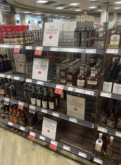 Photo of booze store shelf with emoty shelves where American product used to be, with sign saying Buy Canadian Instead