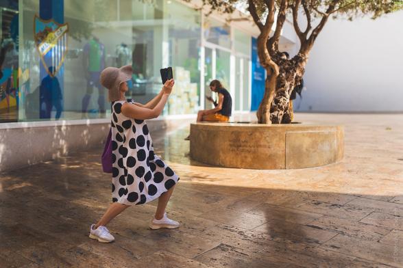 Street photography: Woman in polka dot dress and sun hat taking photo with smartphone outside storefront in Málaga, with decorative tree planter and another person sitting in background.