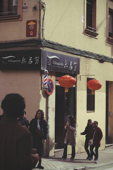 Esquina de una calle con una barbería con farolillos chinos y una pieza de street art con un mosaico de un dibujo con un personaje con ropa tradicional china