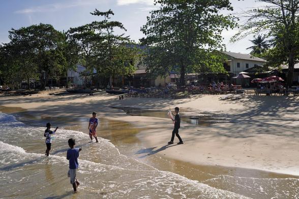 Strand, vier Kinder in Bewegung im flachen Wasser, einer hat einen Drachen in der Hand. Dahinter im Schatten von Bäumen Stühle, Tische und Sonnenschirme, dahinter Häuser.