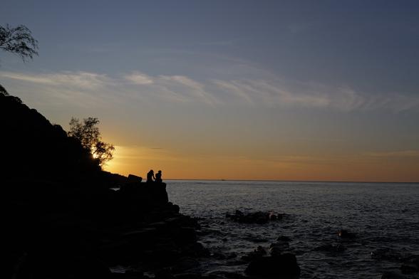 Sonnenuntergang an der Küste, zwei Menschen (als Silhouette) sitzen auf einem Felsen