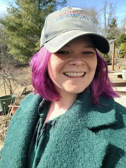 A woman in a hat smiling at the camera. Her hair is short and bright pink/purple