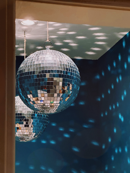 Das Bild zeigt zwei Discokugeln, die an dünnen Seilen von der Decke hängen. Die Spiegelplättchen der Kugeln reflektieren das Licht, wodurch viele kleine Lichtpunkte an die dunkelblauen Wände und die Decke geworfen werden. Eine Lichtquelle oben links sorgt für die Beleuchtung. Die Perspektive ist leicht durch einen Türrahmen oder eine andere Begrenzung eingerahmt, was einen Blick in den Raum ermöglicht.