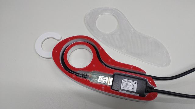 A shoesole-shaped 3D printed part housing a USB plug plugged into a USB extender cable. The cable of the plug is guided in a loop so that two cable ends exit the thing at the right hand side. Everything sits snugly inside groobes so nothing moves. A transparent cap lies next to it. The housing has a small flat hook on the top.