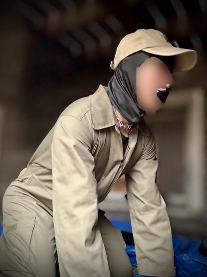 A close-up photo showing a black gag pokes out from the workhorse’s mouth, while a metallic chain collar peeks out at the neck, hinting at a fetish or kink aesthetic. The dynamic pose, with the figure bent forward slightly, suggests movement or action. The blurred background effectively focuses the viewer’s attention on the subject’s silhouette. The combination of tactical/workwear styling with fetish elements creates an arresting visual that skillfully bridges the gap between practical and pony play sensibilities.
