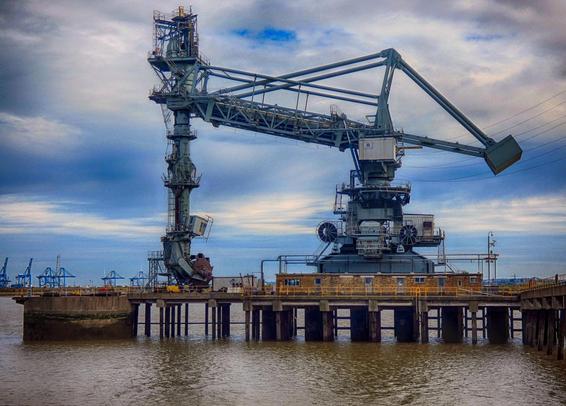 A very large industral machine stands on a wharf, it appears to be like a crane but with a huge hand reaching down to the wharf, and something in that hand.