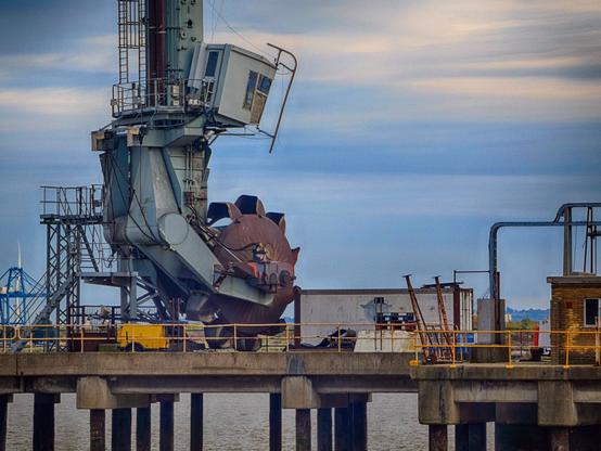 A very large industral machine stands on a wharf, the part of it in shot is like huge hand, and with a circular multi-scooped thing cradled in that multi-storey hand.