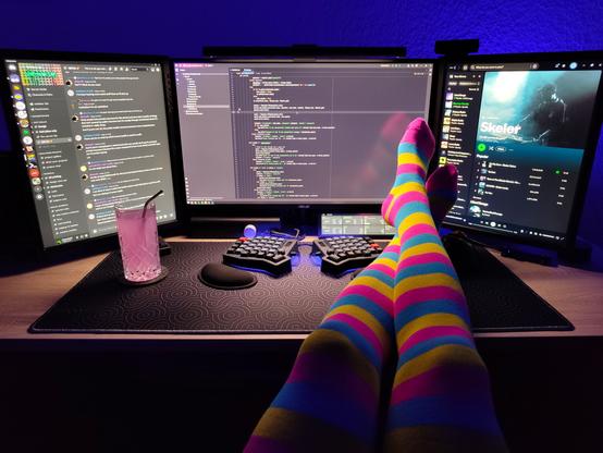 Photo of my desk with three computer monitors on it.
The wall behind the monitors is lit up with blue light.
My legs in pink, yellow and blue striped thigh highs are resting on the desk next to my keyboard.
On the left side of the desk is a glass filled with pink lemonade.