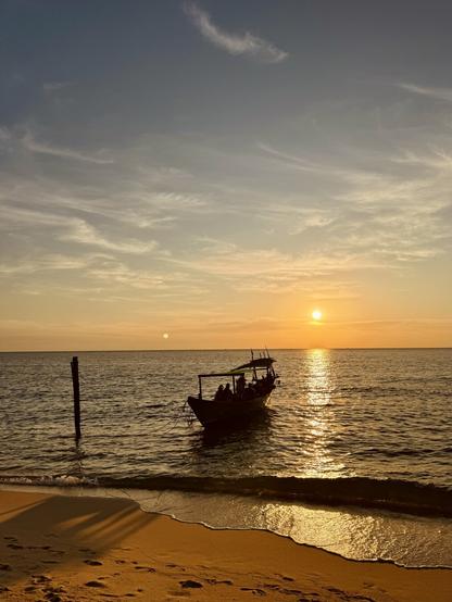 Ein Boot liegt am Strand im Wasser, dahinter die untergehende Sonne. Das Boot wirft einen langen Schatten auf den Sand.