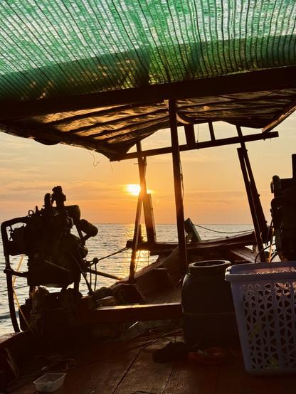 Blick vom Holzboot aus auf das Meer und den Sonnenuntergang. Silhouettenhaft sieht man einen großen Motor und die Balken, die ein Sonnendach tragen.