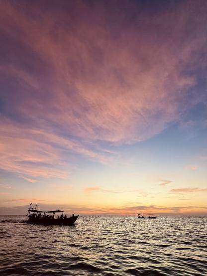 Zwei fahrende Boote auf dem Meer in unterschiedlicher Entfernung. Kurz nach Sonnenuntergang, über den Himmel erstrecken sich rosa-lila-farbene Schleierwolken.