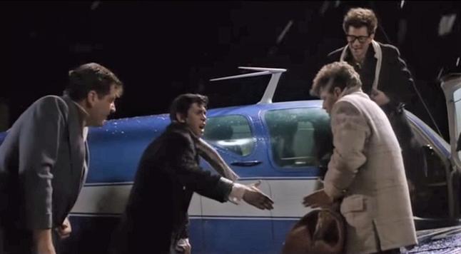 Four young men in 50s clothing and jackets hurry to put their bags and belongings on a small blue plane