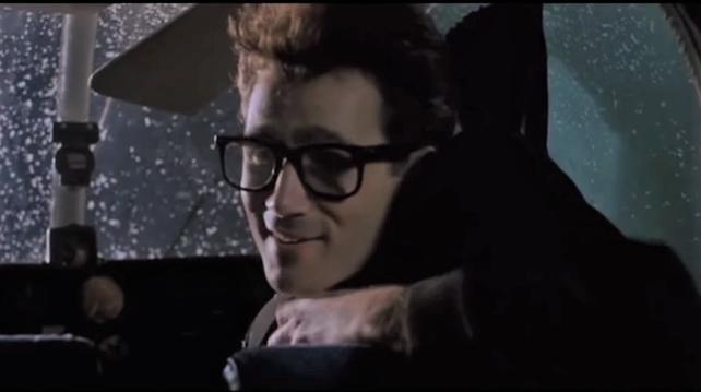 A young man wearing big thick-rimmed glasses turns from the front seat of a plane to smile at someone behind him
