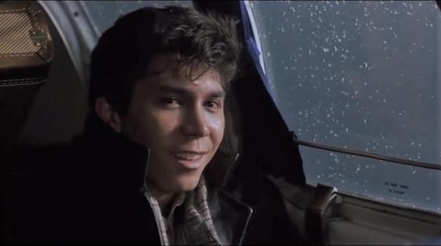 A young man wearing a cool jacket leans against a plane window and smiles at the camera