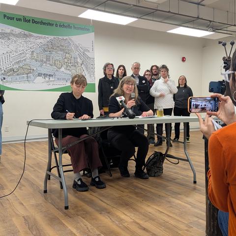 Deux personnes sont assises à une table pliante, l'une d'entre elle parle dans un micro. On voit un autre micro placé devant elle. En arrière, plusieurs personnes se tiennent debout. Au centre, une bannière montrant une image de l'îlot Dorchester et une mention "Pour un îlot Dorchester à échelle humaine."