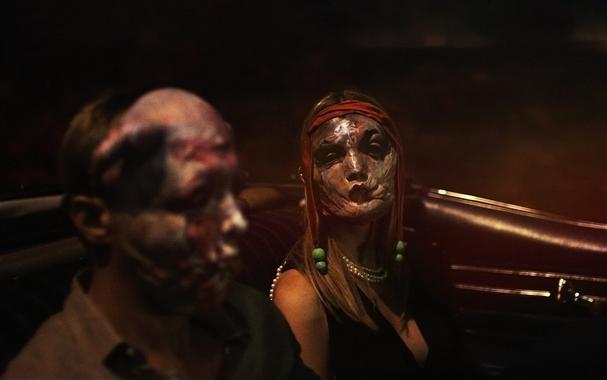 An image from the horror film Infinity Pool, showing a man and a woman sitting in an open top car, both wearing grotesque face masks