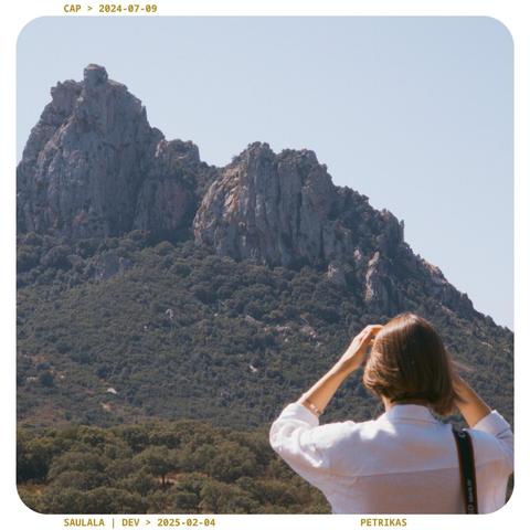 A person in foreground fixing their hair and looking at some rocky mountains in the foreground. Very sunny, hot atmosphere.