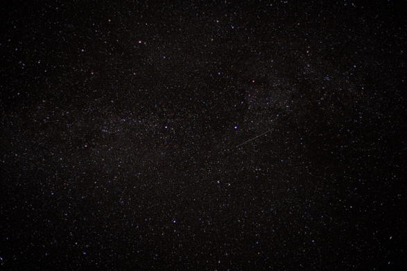 A photograph of the starry night sky taken at Hess Nature Preserve near Presque Isle County, Michigan, in October 2024. The image showcases a densely packed expanse of stars, scattered across the dark sky. A faint streak, likely a meteor or satellite, cuts through the stars, adding a dynamic touch to the scene.