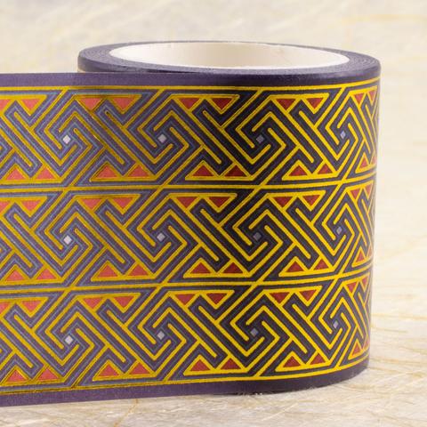 A roll of wide wash tape (45mm) stands on its end with the tape unspooling to the left to show the design, a Celtic key/maze pattern on a deep purple background is outlined in gold with red and pale blue accents.