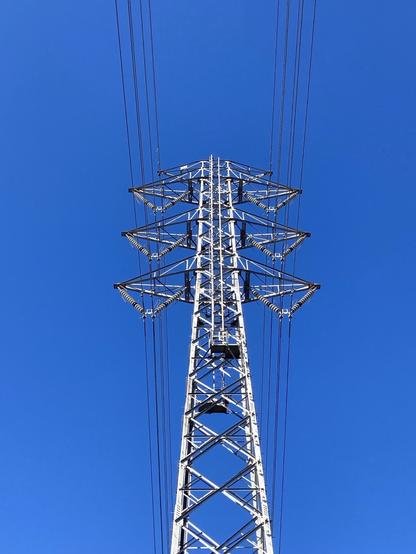 A tall metal power transmission tower against a clear blue sky, with high-voltage wires extending from its top.
