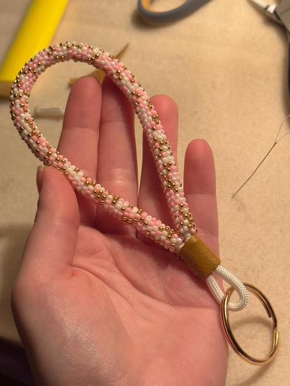 A hand holding a mini bead wrapped keychain in pale pink, white, and gold, with a gold split ring