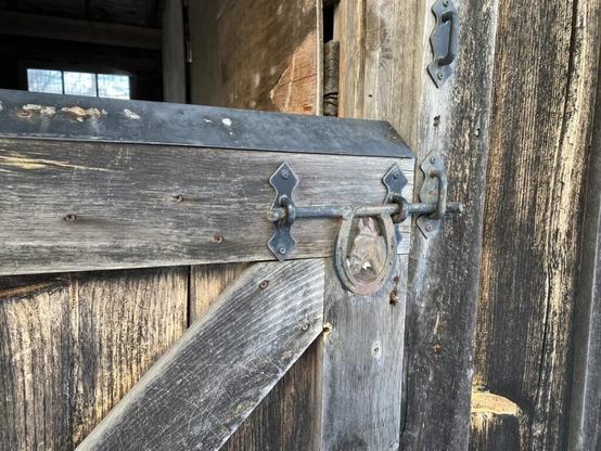 A lower half of an unpainted, weathered barn door that is latched closed with an iron latch. The latch is designed decoratively with a horseshoe, and inside the open space of the horseshoe is a horse's head.