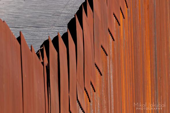 An abstract "arty" shot of the top of the U.S.-Mexico border fence. It appears as a series of pointy, rusty metal plates, like some sort of metallic dragon's-back plates atop vertical corrugations formed by telephoto compression of the 30' tall posts that make up the fence. The border fence road can be seen "above" the fence, though it's really an artifact of the telephoto lens and the angle of view. The road surface appears as black and white corduroy stripes: shadows from the low sun shining through the fence.
