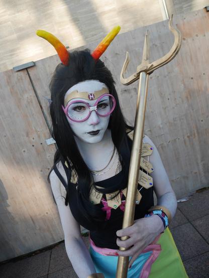 Cosplay of the character Feferi Peixes of the comic Homestuck. Feferi has grey skin, black long hair and curved orange/yellow horns on her head. The cosplayer is standing in front of a wooden wall and holding Feferis signature weapon, a golden trident.
