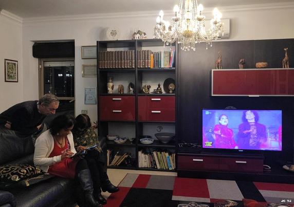 Farbfoto, das ein Wohnzimmer mit einem großen Regal mit Büchern, Deko und Fernseher, Teppich, Bildern, einer Tür nach draußen und drei Personen auf einem Sofa ganz links zeigt. Alles wird bestrahlt durch einen Kronleuchter oben rechts von der Bildmitte. Der Fernseher zeigt ein Bild einer Gala.