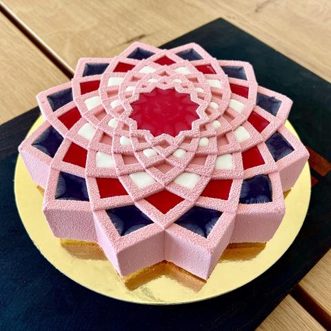 a 12-point star shaped cake sprayed in light pink cocoa butter velvet, with concentric little pockets filled with 3 colors of mirror glaze (red, purple, and white). The cake is placed on a golden circular cake board and on a rectangle dark exotic wood cutting board.