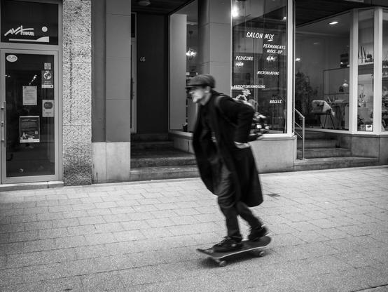A black-and-white street photo taken in Blankenberge, Belgium, showing a skater in a long black coat and a cap moving through a shopping street.