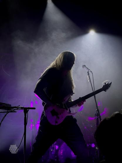 A long-haired person with long hair is silhouetted playing a light-colored electric guitar on a dark stage with purple lighting. A microphone is on a stand to the left of the guitarist. There is a keyboard or synthesizer in the background to the left, partially obscured by smoke or haze. Part of a drum kit is visible in the bottom right. The overall scene is dimly lit, with spotlights highlighting the guitarist.