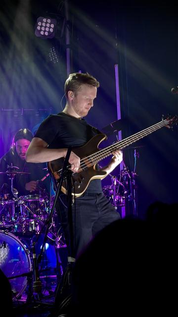 A man with short, light brown hair plays a bass guitar on a stage lit with purple and blue stage lighting. He's wearing a black t-shirt and dark pants. Behind him, partially obscured, is a drummer sitting at a drum kit. A stage light with multiple bulbs is visible in the upper left corner.
