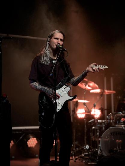 A long-haired man with tattoos on his arms is singing into a microphone while holding only the corpus and the head of an electric guitar on stage. He is wearing a dark shirt and dark pants. A drum kit is visible in the background, out of focus. There is stage lighting illuminating the scene.
