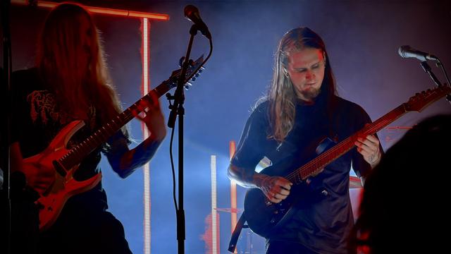 Two long-haired men play electric guitars on a stage lit with blue and red vertical lights. The guitarist on the left plays a light-colored guitar and the guitarist on the right plays a dark-colored guitar. Microphones are visible on stands in front of each guitarist.