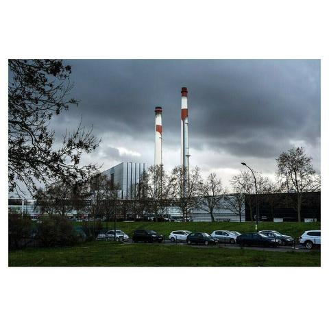 Un paysage industriel se déploie sous un ciel lourd et menaçant, chargé de nuages sombres. Deux hautes cheminées, blanches avec des anneaux rouges, se dressent au centre de l’image, dominant l’horizon. Elles appartiennent à un grand bâtiment moderne en verre et métal, partiellement dissimulé par une rangée d’arbres dénudés, suggérant une saison hivernale.

Au premier plan, une étendue de pelouse verte contraste avec la grisaille ambiante. Une route bordée de voitures stationnées traverse la scène, séparant l’espace naturel de l’installation industrielle. Un lampadaire courbé, placé sur la droite, accentue l’atmosphère urbaine. L’ensemble dégage une sensation de solitude et de froideur, où la présence humaine est suggérée mais absente.