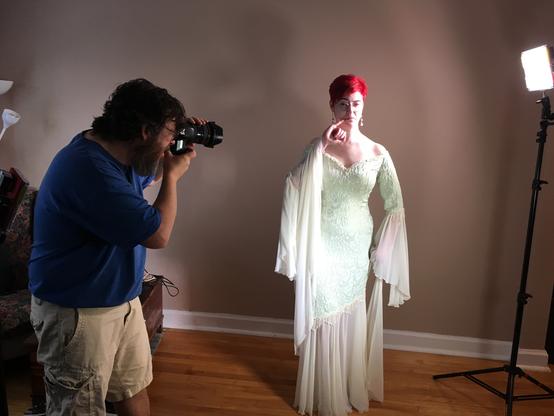 A man with a beard in a blue shirt and khaki shorts takes a photo of a red headed model in a green brocade and ivory chiffon bridal gown. She has a quirky look on her bespectacled face with one pinky finger at her lips.
