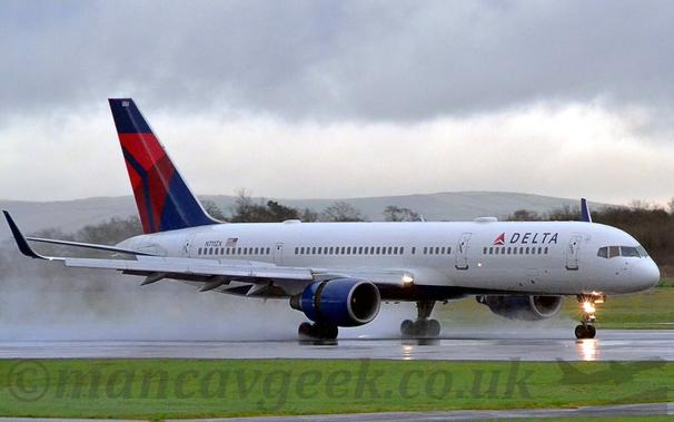 Side view of a white, twin engined jet airliner slowing down as it moves from left to right along a wet, black runway.
Spray is being kicked up by the wheels and the exhaust from the engines.
The plane has black "Delta" titles on the upper forward fuselage, with the registration "N711ZX" on the upper rear fuselage, next to an American flag.
The engine pods slung under the wings have the rear section pushed back, allowing the thrust reversers to push air forwards to slow the plane down.
There is a bright light mounted on the nose wheel, pointing forwards.
Green grass fills the bottom of the frame in the foreground, with more grass on the other side of the runway, leading up to trees in the distance.
In the far distance, rolling hills give way to stormy grey clouds.