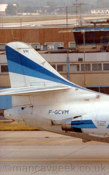 Close up of the white rear fuselage and tail of a twin engined jet airliner facing to the right with the engines mounted on the sides of the rear fuselage.
The tail has a two-tone blue running from the lower front edge to the top rear, getting wider as it goes.
A similar stripe runs along the engine pods, with a white section in the middle with a winged logo with "Air Provence" title above it.
The registration "F-GCVM" is on the upper rear fuselage, just aft of the engines, in blue.
A wide building in shades of brown stretches across the background, with trees and low buildings in the distance.